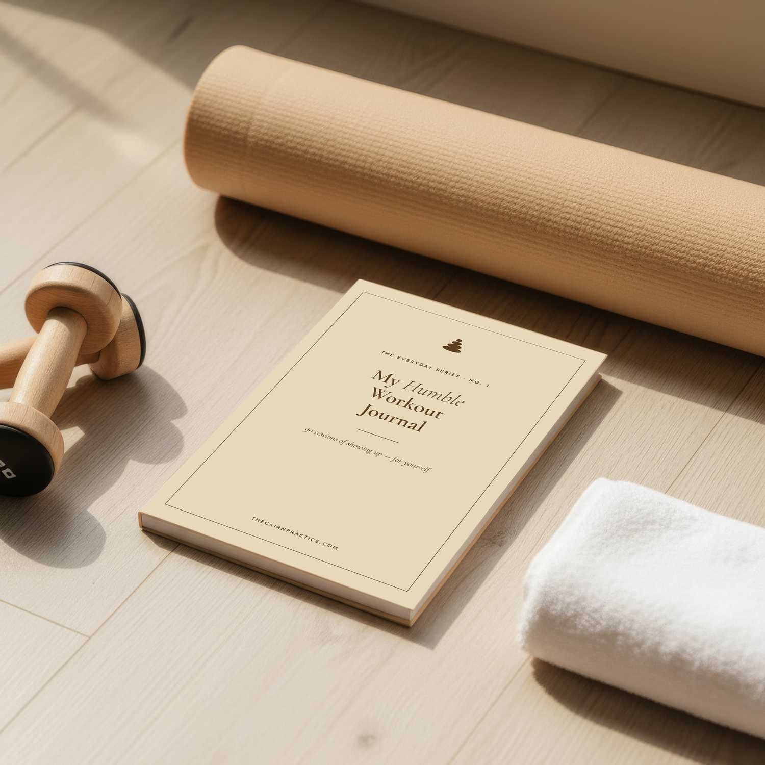 A styled flat-lay of a beige journal titled 'My Humble Workout Journal' on a light wood floor, alongside a rolled tan exercise mat, a wooden dumbbell, and a white towel.