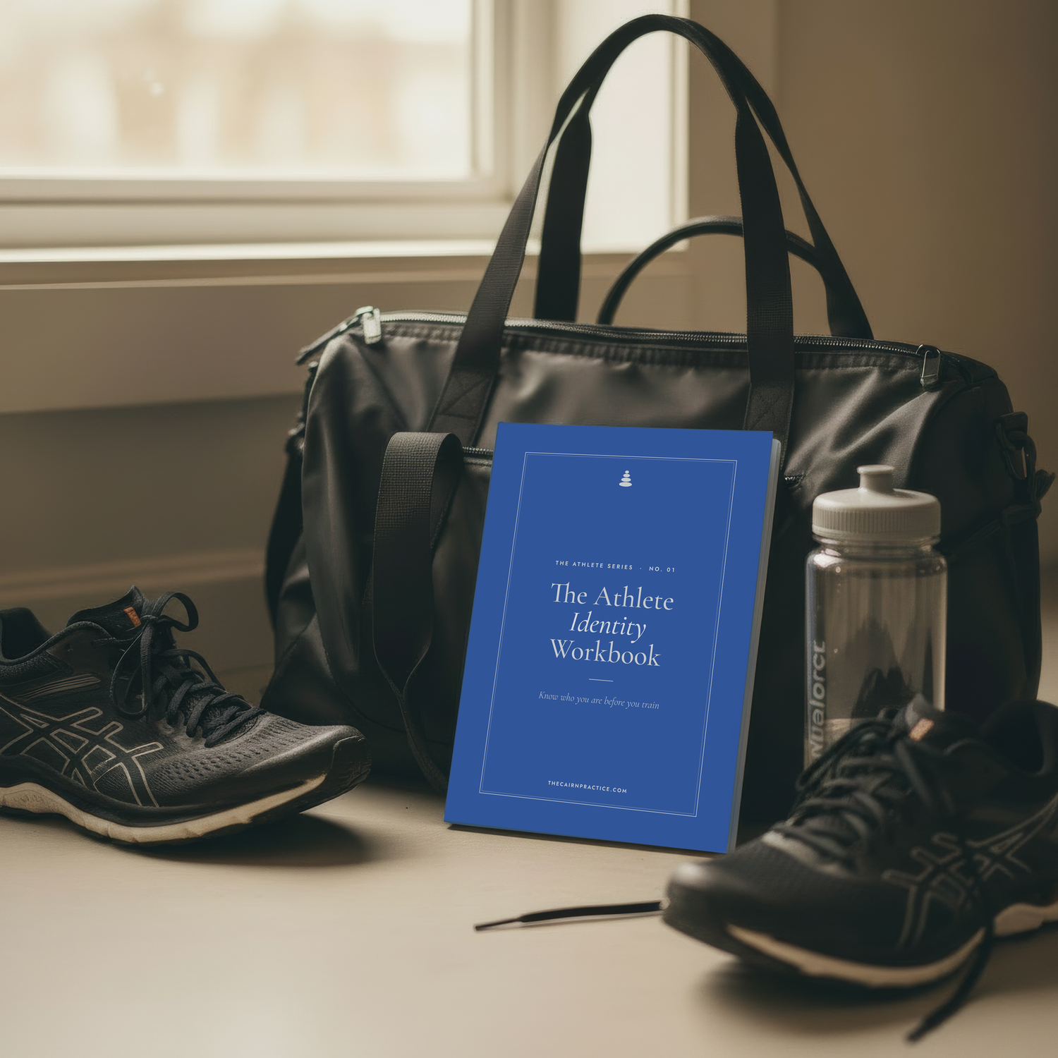 A blue booklet titled 'The Athlete Identity Workbook' stands in front of a black duffel bag on a tabletop, surrounded by athletic shoes and a clear water bottle in a softly lit indoor setting.
