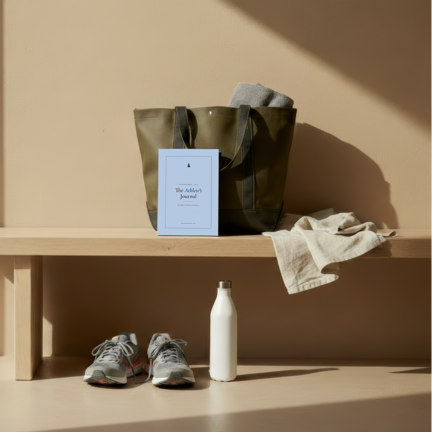 An olive green canvas tote bag sits on a light wood bench against a warm beige wall, with a light blue journal, a folded gray blanket, and a cream linen cloth beside it. A pair of gray sneakers and a white bottle are placed on the floor below.