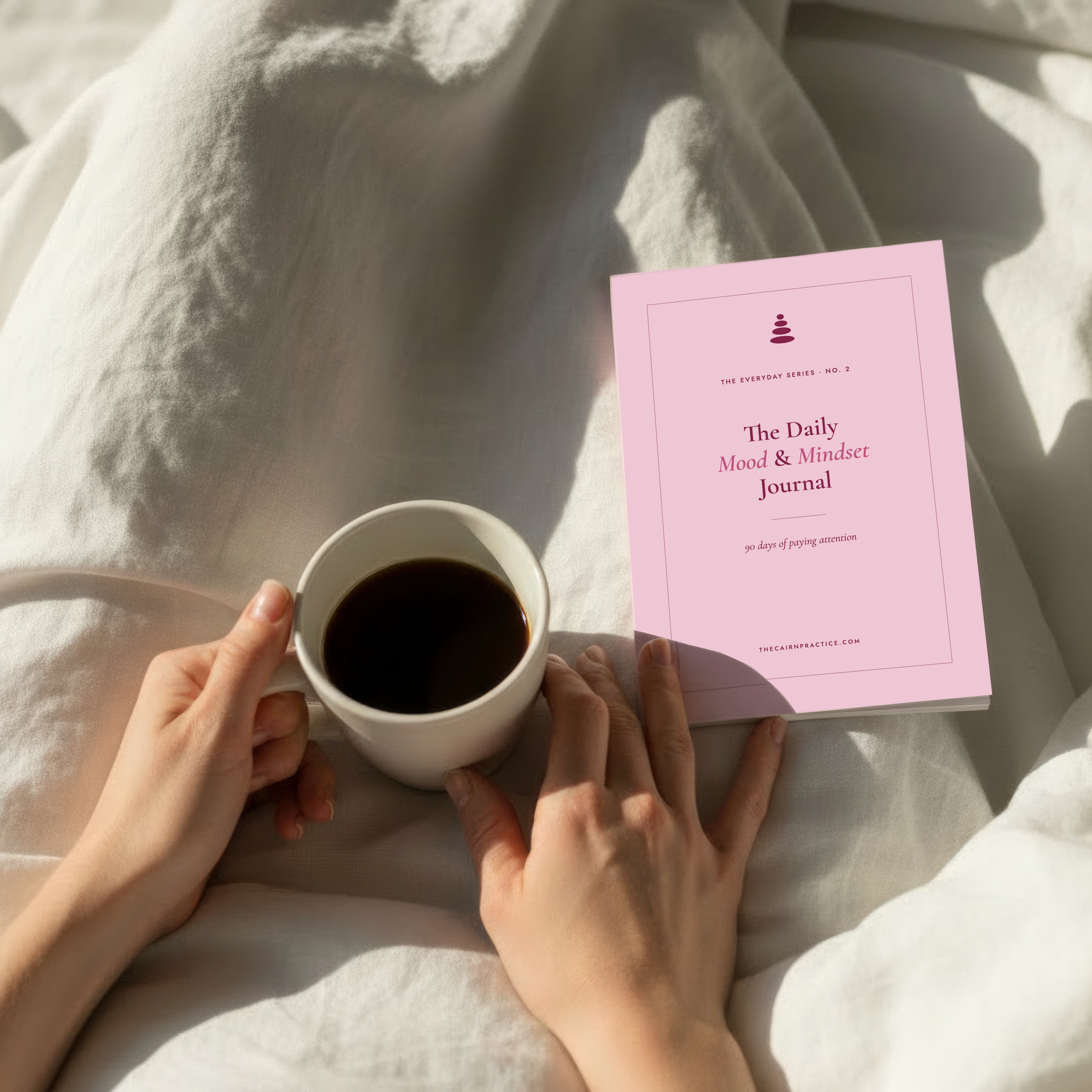 Person holding a cup of coffee next to a pink journal on a soft surface
