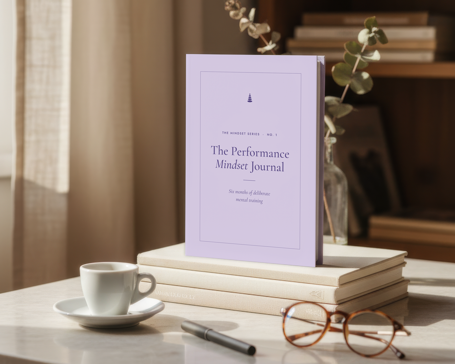 A lavender hardcover journal titled 'The Performance Mindset Journal' stands upright on a stack of books on a table, with a white cup, pen, eyeglasses, and a vase in a softly lit indoor setting.